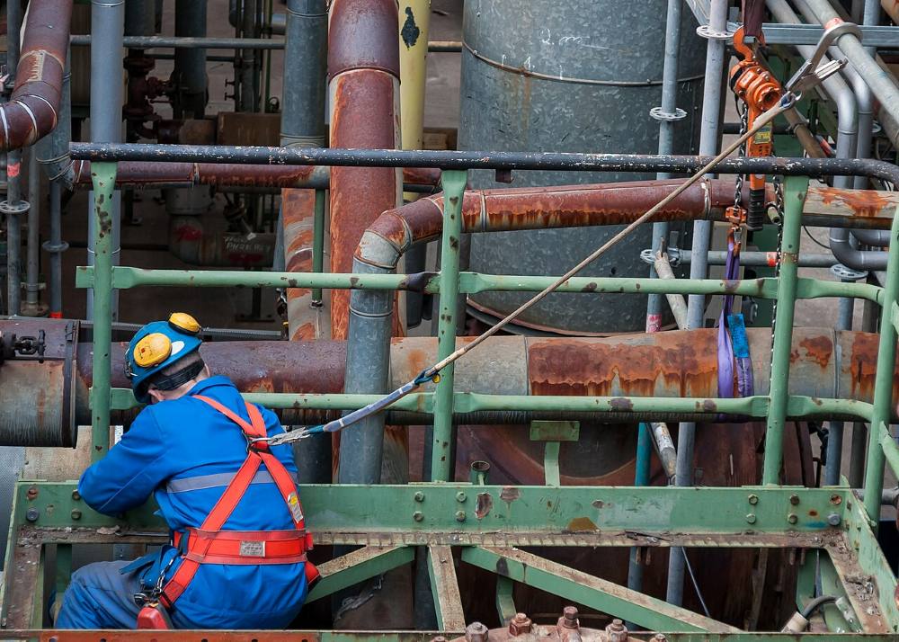 Trabajador con arnés de seguridad y línea de vida anclada, sobre una plataforma entre cañerías industriales.